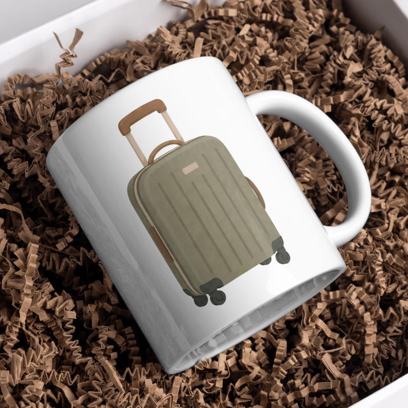 May include: White ceramic mug featuring a green suitcase illustration with brown accents. The suitcase has a retractable handle and wheels. The mug is set against a background of brown packing material.