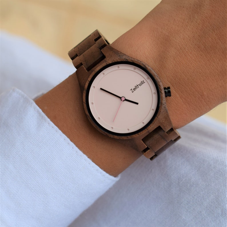 May include: A brown wooden wristwatch with a pink face and black hands. The watch face has the word "Zeitholz" printed on it.