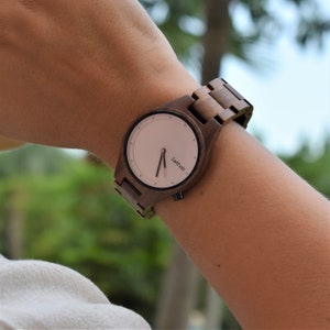 May include: A brown wooden wristwatch with a pink face and black hands. The watch face has the word "Zeit holz" printed on it.