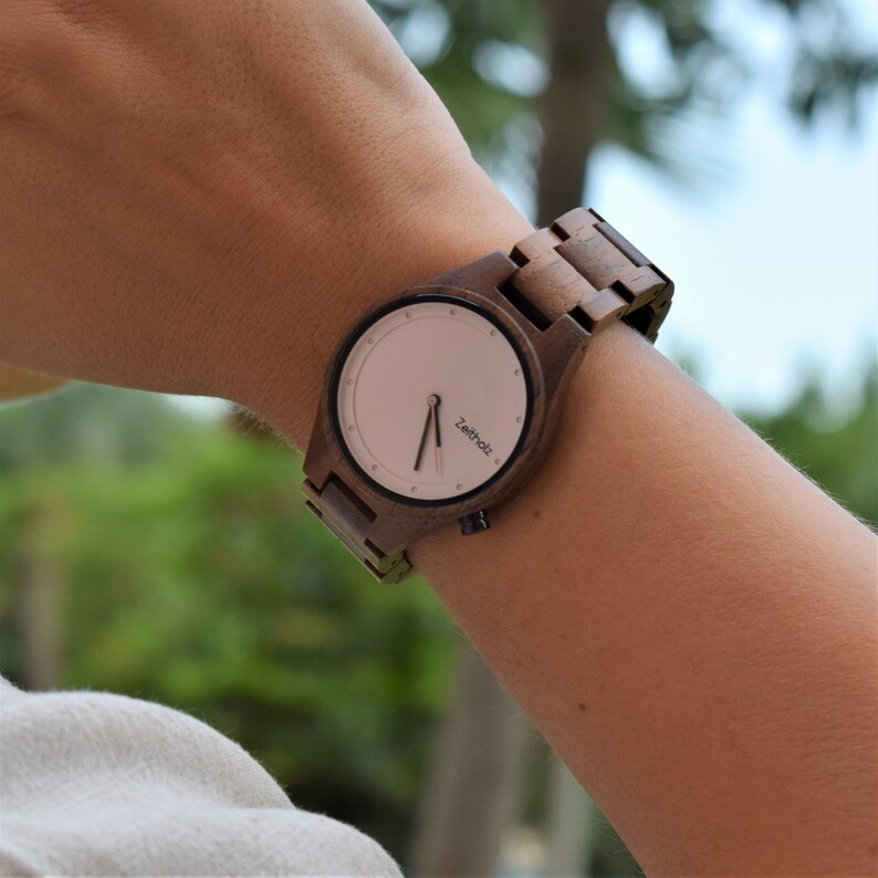 May include: A brown wooden wristwatch with a pink face and black hands. The watch face has the word "Zeit holz" printed on it.