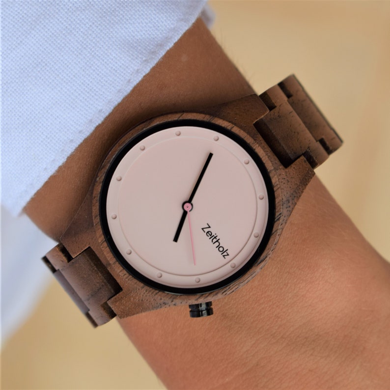 May include: A brown wooden wristwatch with a pink face and black hands. The watch face has the word "Zeitholz" printed on it.