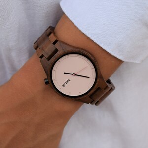 May include: A brown wooden wristwatch with a pink face and black hands. The watch face has the text "Z&Auml;HRLOS" printed on it.