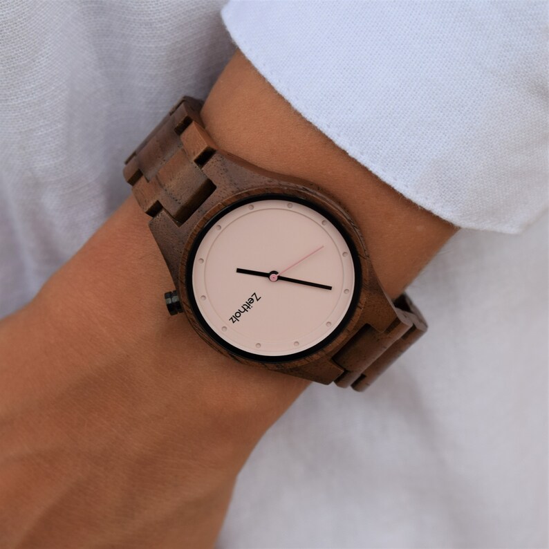 May include: A brown wooden wristwatch with a pink face and black hands. The watch face has the text "Z&Auml;HRLOS" printed on it.