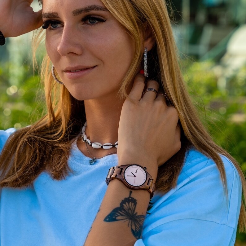 May include: A woman wearing a light blue shirt with a wooden watch on her wrist. The watch has a white face with black hands and a brown wooden band. The woman has a butterfly tattoo on her wrist.