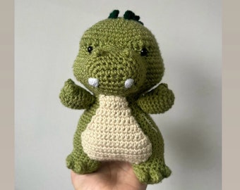 May include: A green crocheted dinosaur toy with white teeth and black eyes. The dinosaur is standing on its two feet and has its arms outstretched.