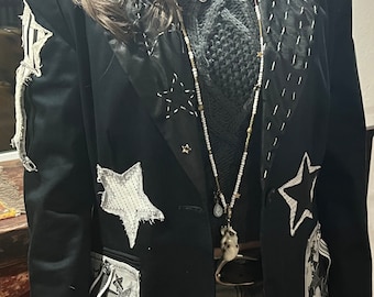 May include: A black blazer with a variety of star-shaped patches sewn on. The blazer has a white collar and a silver chain necklace with a small eye charm.