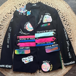 May include: Black blazer with colorful patches and handwritten text. Patches include tie-dye designs, rainbows, and phrases like "Fix You," "People of Pride," and "Magic." The blazer has a classic tailored fit.