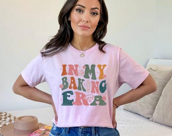 May include: A light pink t-shirt with the text "IN MY BAKING ERA" in a retro font. The text includes images of a whisk, rolling pin, and chef's hat. The shirt is being worn by a person.