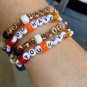Könnte beinhalten: Vier Armbänder mit weißen Perlen und schwarzen Buchstaben, die jeweils "YOU HAVE" buchstabieren. Die Perlen sind mit goldenen und orangefarbenen Perlen durchsetzt.