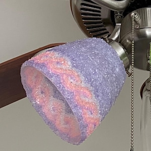 Könnte beinhalten: Ein Deckenventilator mit dekorativer Leuchte. Die Leuchte hat einen lavendelfarbenen, strukturierten Schirm mit einem rosa-orangenen Muster. Die Lüfterflügel sind dunkelbraun.