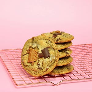 May include: A stack of six round golden-brown cookies featuring visible chunks of dark chocolate and pieces of light brown biscuit. The cookies are resting on a metallic rose-gold wire cooling rack with a grid pattern.