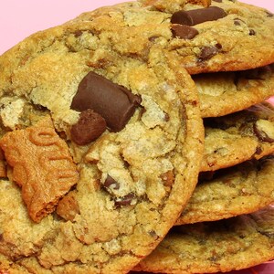 May include: A stack of golden-brown baked cookies featuring large dark chocolate chunks and a piece of a textured, light brown spiced biscuit embedded in the surface. The cookies have a rough, uneven surface with visible chocolate pieces.