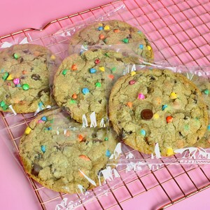 May include: Five chocolate chip cookies with colourful sprinkles, individually wrapped in clear plastic bags, on a pink wire rack.