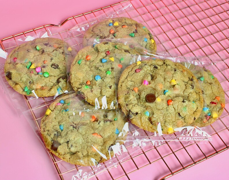 May include: Five chocolate chip cookies with colourful sprinkles, individually wrapped in clear plastic bags, on a pink wire rack.