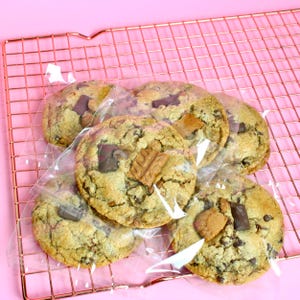 May include: Five round, golden-brown cookies with visible dark chocolate chunks and light brown textured pieces, each individually wrapped in clear plastic packaging and arranged on a copper-colored metal wire cooling rack.