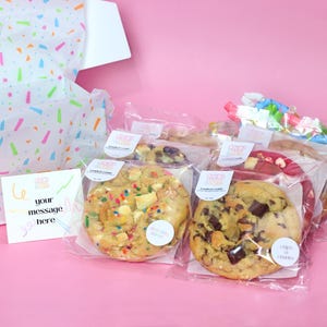 May include: Four individually packaged cookies in clear plastic bags. The cookies are chocolate chip, oatmeal raisin, white chocolate chip, and red velvet. Each bag has a sticker with the text "Cookies & Cream" and a small circle with the name of the cookie flavor.