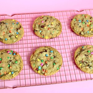 May include: Six chocolate chip cookies with colourful sprinkles on a copper wire cooling rack. The cookies are on a pink background.