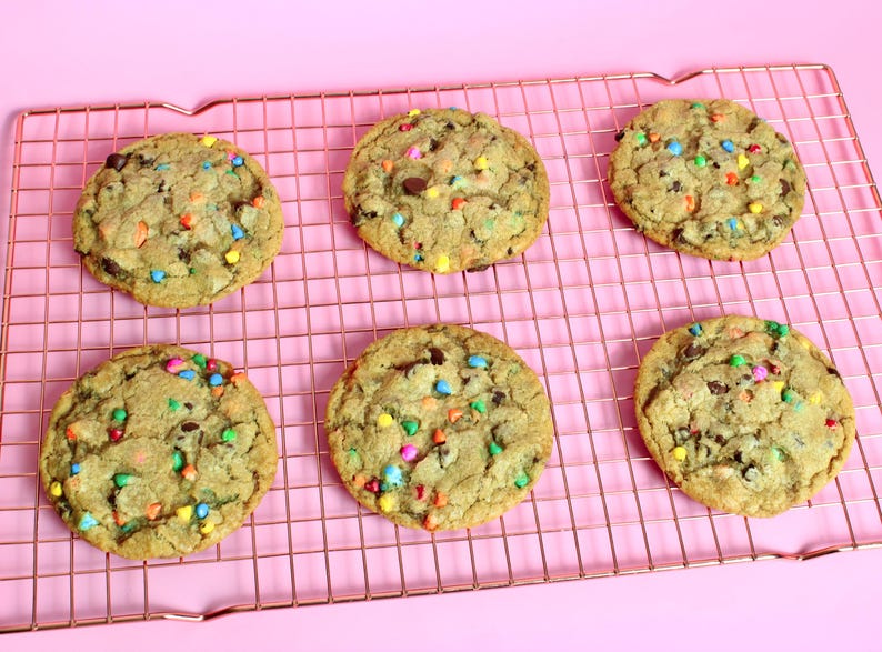 May include: Six chocolate chip cookies with colourful sprinkles on a copper wire cooling rack. The cookies are on a pink background.