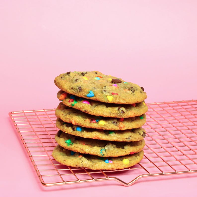 May include: A stack of six chocolate chip cookies with colourful sprinkles on a gold wire cooling rack. The cookies are on a pink background.