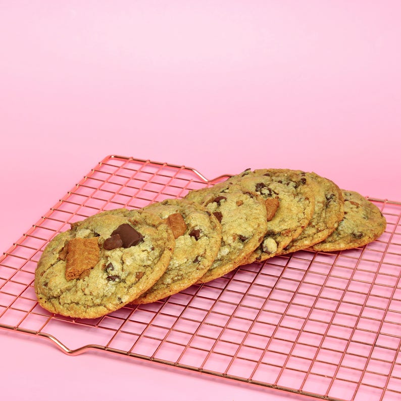 May include: A row of five round, golden-brown chocolate chip cookies with visible chunks of chocolate and pieces of golden honeycomb toffee, arranged in a diagonal line on a copper-colored metal wire cooling rack against a solid pink background.
