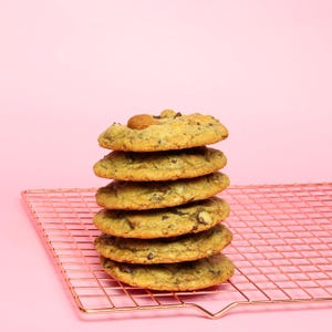 May include: A vertical stack of six golden-brown chocolate chip cookies resting on a rose gold metal wire cooling rack. Each cookie features visible dark chocolate chunks and a slightly uneven, baked texture, creating a rustic appearance.