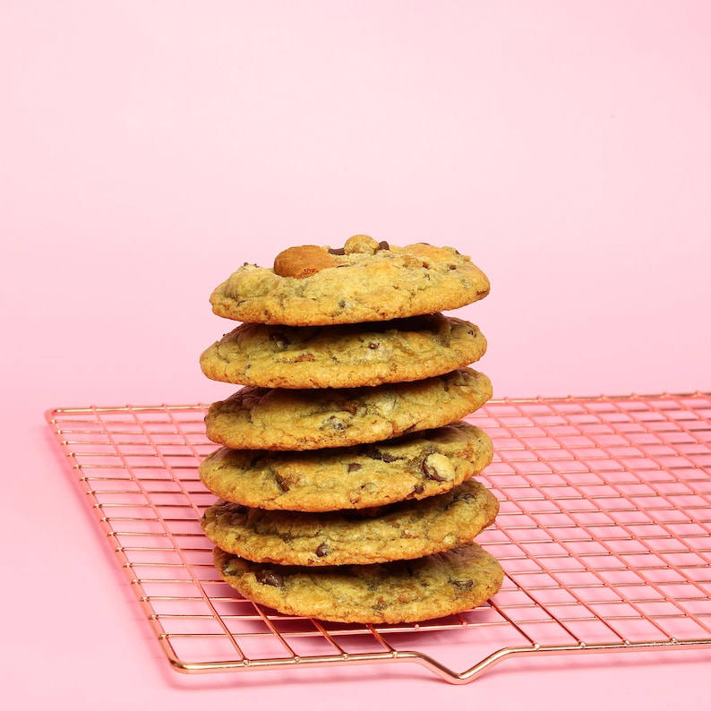 May include: A vertical stack of six golden-brown chocolate chip cookies resting on a rose gold metal wire cooling rack. Each cookie features visible dark chocolate chunks and a slightly uneven, baked texture, creating a rustic appearance.