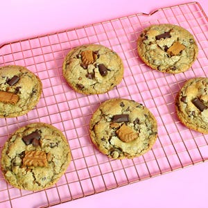 May include: Five round, golden-brown cookies arranged on a copper-colored wire cooling rack. Each cookie features visible dark chocolate chunks and pieces of light brown, textured biscuit embedded in the dough, creating a varied surface.