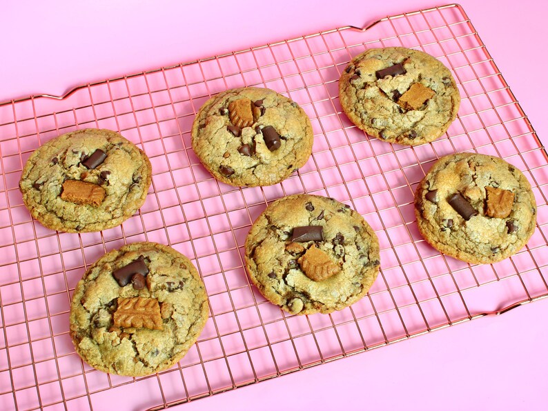 May include: Five round, golden-brown cookies arranged on a copper-colored wire cooling rack. Each cookie features visible dark chocolate chunks and pieces of light brown, textured biscuit embedded in the dough, creating a varied surface.