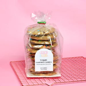 May include: A clear plastic bag containing a stack of 12 vegan gourmet cookies. The cookies are a light brown colour with colourful sprinkles. The bag has a label that reads "GRUMB & SWIRL Vegan Gourmet Cookies deliciously thin & flavorful 12 Cookies (2 oz each)"