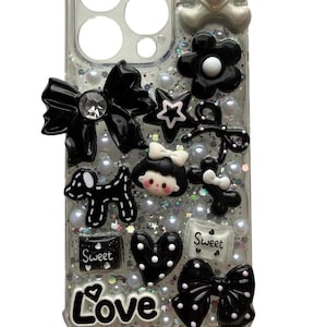Può includere: Custodia per telefono trasparente decorata con fiocchi neri, un orsacchiotto, fiori, stelle e un cuore. La custodia ha uno sfondo glitterato con accenti di perle e la parola "Love". Il testo "Glitz Charm Lab" è visibile.