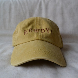 Peut inclure: Une casquette de baseball jaune avec le mot "HOWDY!" brodé en marron sur le devant.