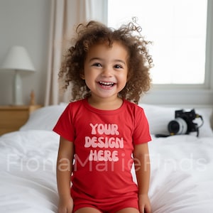 May include: A young child wearing a red short-sleeved shirt with the text "YOUR DESIGN HERE" in pink letters. The child is sitting on a white bed and smiling.