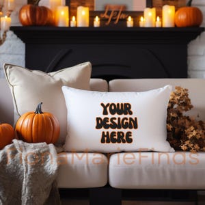 Pode incluir: Almofada retangular branca com o texto "YOUR DESIGN HERE" em preto e laranja. A almofada está em um sofá bege, com abóboras e velas ao fundo. Um item decorativo com tema de outono.