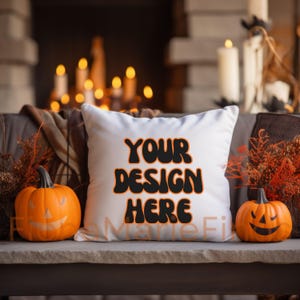 Op de afbeelding: Wit vierkant kussen met de tekst "YOUR DESIGN HERE" in zwart met oranje omlijning. Twee uitgesneden oranje pompoenen flankeren het kussen. De scène is gecreëerd met kaarsen en herfstbladeren, wat een Halloween-thema creëert.