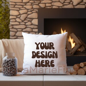 Puede incluir: Almohada blanca con el texto "YOUR DESIGN HERE" en letras marrones. La almohada está sobre una superficie de madera delante de una chimenea.