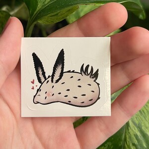 Könnte beinhalten: Ein Aufkleber mit einem Cartoon-Seehase mit weißem Körper, schwarzen Punkten und schwarzspitzigen Ohren. Der Seehase hat ein rosa Gesicht und ist von kleinen rosa Herzen umgeben. Der Aufkleber wird in einer Hand gehalten.