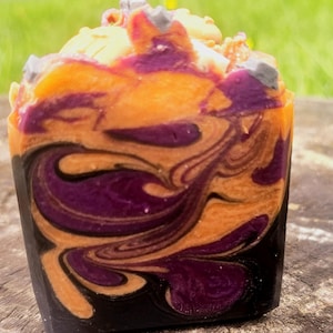 May include: A rectangular bar of soap featuring a marbled swirl pattern of deep purple, bright orange, and black colors. The top of the soap is textured with irregular, light-colored peaks and small grey accents, creating a multi-layered appearance.
