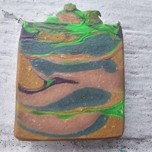 May include: A rectangular bar of handmade soap with a layered design. The soap features swirls of green, teal, gold, and brown colours. The top of the soap has a textured, uneven surface with a green and brown design.