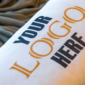 May include: White pillow with the text "YOUR LOGO HERE" in black and gold lettering. The pillow is lying on a wooden surface.