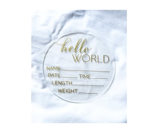 May include: A clear acrylic disc with gold lettering that reads "hello WORLD." Below the text are lines for Name, Date, Time, Length, and Weight. The disc is a baby announcement or keepsake.