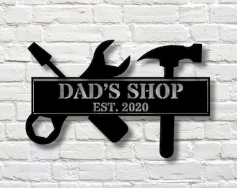 Può includere: Segno in metallo nero con una chiave inglese, un cacciavite e un martello, con il testo "DAD'S SHOP EST. 2020" su sfondo nero.