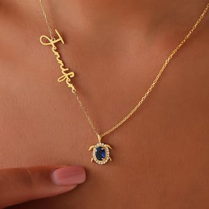 May include: A gold-colored necklace featuring the name "Jennifer" in cursive script. The necklace includes a turtle pendant with a blue gemstone center and a halo of small, clear stones. The chain is delicate and the overall design is elegant.