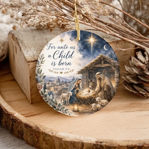 May include: A round ceramic ornament featuring a nativity scene with Mary, Joseph, and the baby Jesus in a manger. The text reads For unto us a Child is born Isaiah 9:6. The design includes a bright star, a stable, sheep, and a town landscape.