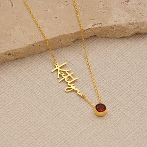 May include: A gold-colored necklace with the name "Katelyn" in a cursive font. The necklace has a small, round, red gemstone pendant. The chain is delicate and the pendant is set in a gold bezel.