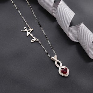 May include: A silver necklace with the name "Amy" in cursive script. The pendant features a red heart-shaped gemstone surrounded by a halo of small, clear stones. A gray ribbon is in the background.