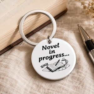 Op de afbeelding: Een ronde, witte sleutelhanger met de zwarte tekst "Novel in progress...". De sleutelhanger heeft een tekening van een open boek, een bril, een pen en een kopje koffie. Een zilveren sleutelring is bevestigd.