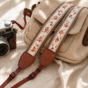 Puede incluir: Una correa de cámara con un estampado floral en tonos melocotón y marrón. La correa se muestra con una bolsa de cámara beige y una cámara de estilo vintage con una funda de cuero marrón. La correa tiene extremos de cuero marrón y correas ajustables rojas.