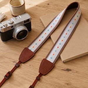 May include: A camera strap featuring a central white fabric band with a repeating pattern of small orange and blue flowers, bordered by brown woven edges and brown leather attachment tabs with triangular stitching and a camera icon embossed.