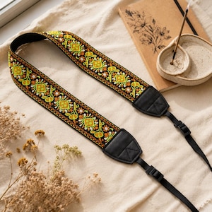 May include: A camera strap featuring a black fabric base with a repeating embroidered floral pattern in shades of orange, yellow, and lime green. The strap includes black leather-like end tabs with stitched detailing and adjustable black plastic buckles.