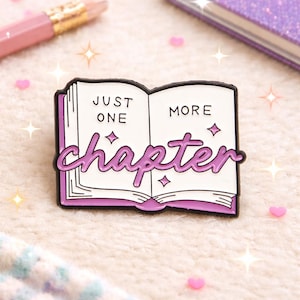 Può includere: Una spilla in smalto viola e bianco a forma di libro aperto. La spilla presenta le parole "JUST ONE MORE chapter" in un font stilizzato. La spilla ha un bordo nero ed è decorata con piccole stelle e diamanti.